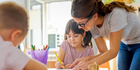 Eine Lehrerin hilft 2 Kindern am Tisch ein Aufgabenblatt auszufüllen