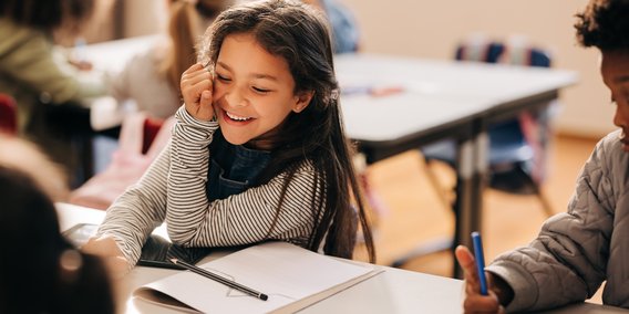 Lachendes Mädchen im Schulzimmer mit Arbeitsheft und Tablet